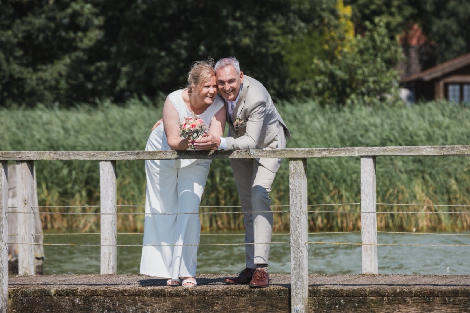 Braut Stefanie und Bräutigam Bernd lehnen lächelnd an einem Holzgeländer auf einem Steg am See